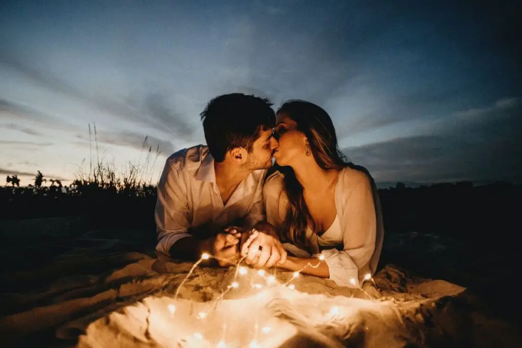 A couple shares a romantic kiss on the beach, illuminated by the glow of sparklers in the evening sky.